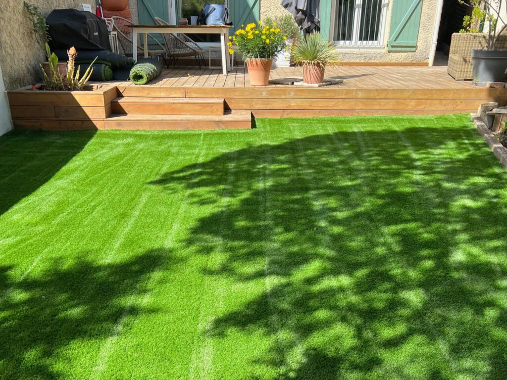 Image principale (jardin avec terrasse bois et façade aux volets bleu-vert) : pose-pelouse-synthetique-bentgrass-52mm-jardin-terrasse-bois-gard-yards